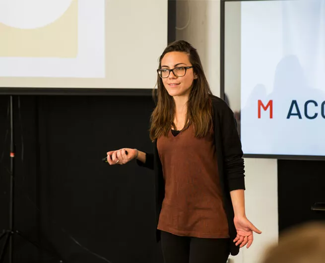 Femme debout très certainement face à une assemblée. Elle tient dans sa main une télécommande pour gérer elle même sa présentation. Cette photo est utilisée pour représenter l'atelier pour tester ton pitch (et enfin être à l’aise pour parler de ton activité)